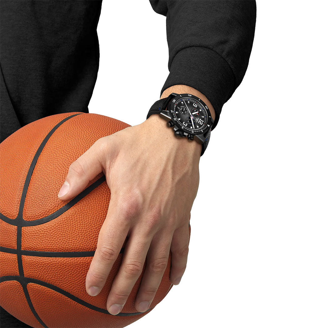 Against a white background, a person in a black long-sleeve shirt holds an orange basketball with their right hand while wearing the Tissot Supersport NBA Special Edition Quartz 45.5mm Watch.