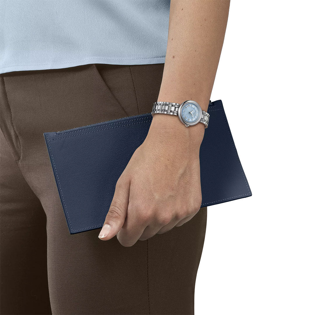 A person wearing brown pants, a light blue shirt, and a Tissot Bellissima Small Lady Quartz 26mm Watch with a stainless steel case holds a navy blue rectangular envelope or wallet at their side.