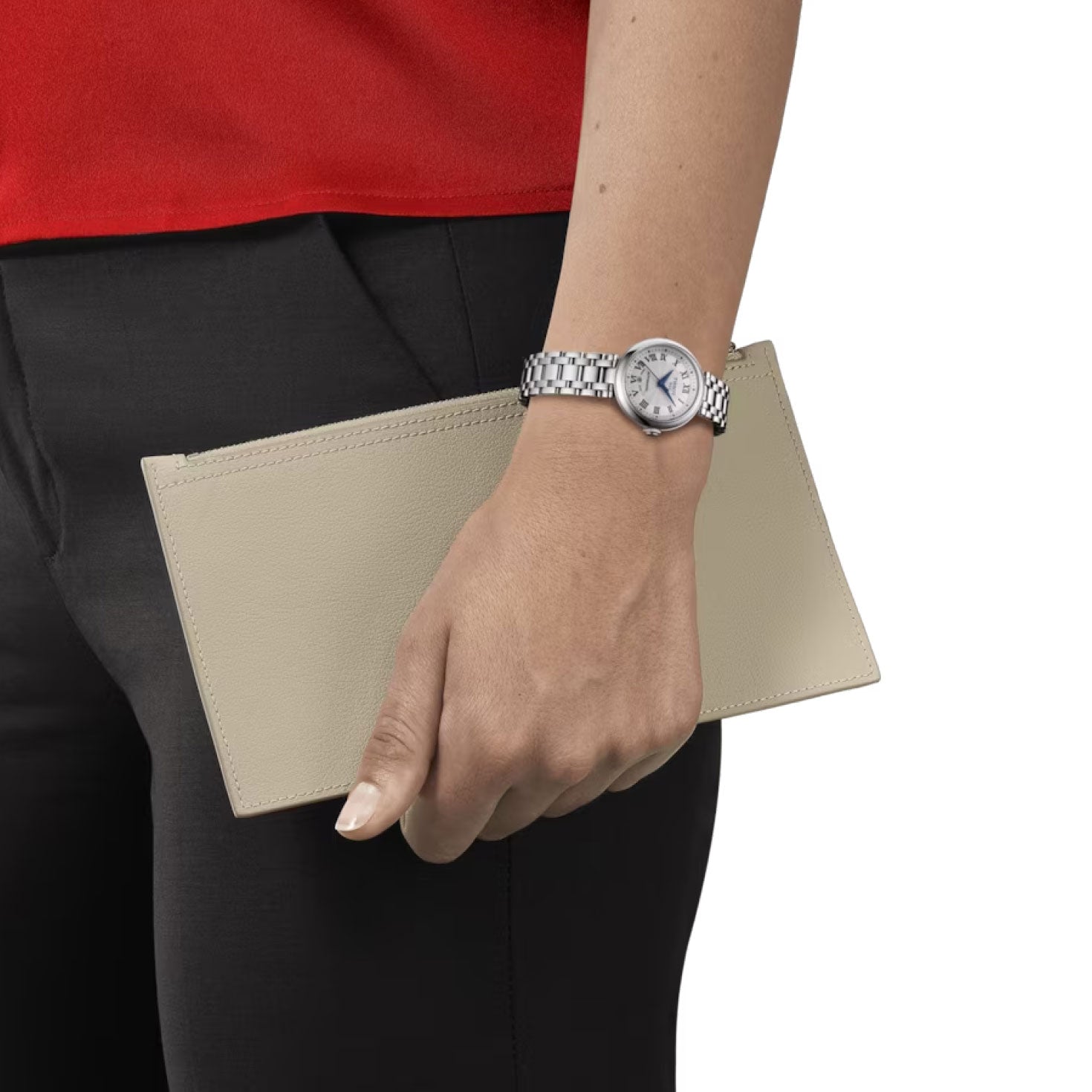 An individual, dressed in a red top and black pants, is seen against a white background wearing a Tissot Bellissima Automatic 29mm watch with Swiss Automatic Movement while holding a beige wallet.