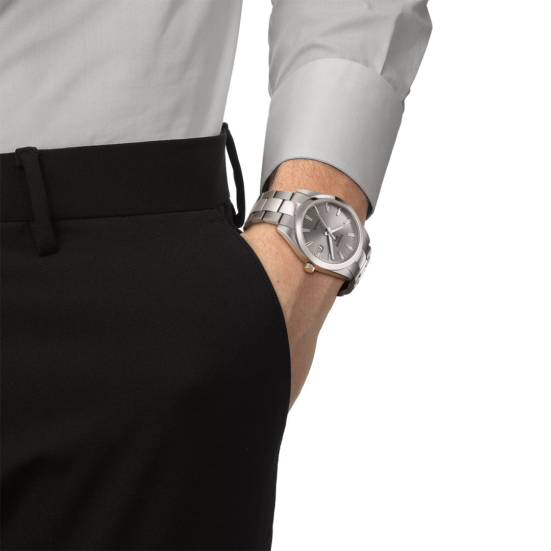 A person in black pants and a light gray dress shirt, with their left hand in their pocket, is wearing a Tissot Gentleman Titanium Quartz 40mm Watch by Tissot on their wrist.