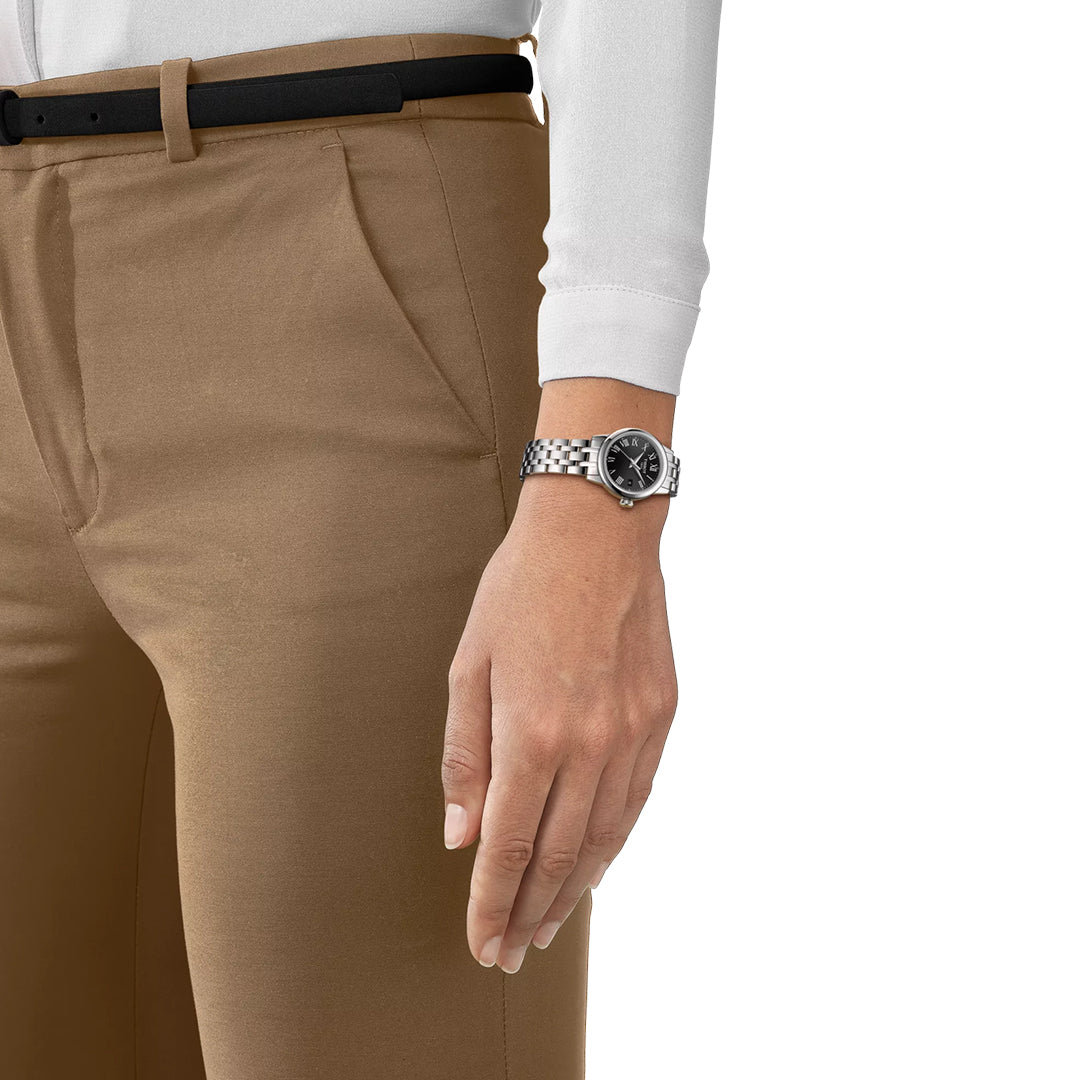 A person in tan pants and a white long-sleeve shirt with a black belt showcases the Tissot Classic Dream Lady Quartz 28mm watch, featuring a dark dial, on their left wrist. Only the lower torso and left arm are visible.