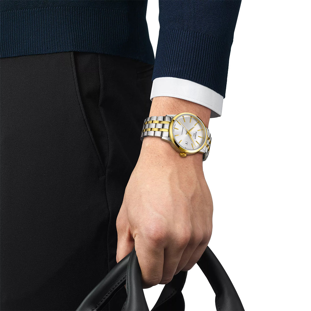 A person in a dark blue sweater and black pants holds black handles, wearing the Tissot Classic Dream Swissmatic 42mm Watch with a white face and yellow gold PVD coating on their wrist.