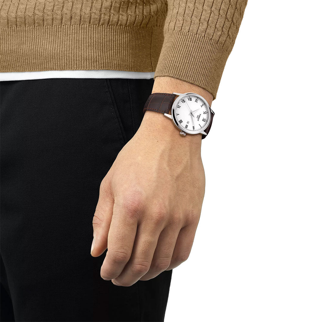 A person in a tan sweater and black pants showcases the Tissot Classic Dream Quartz 42mm Watch, featuring a brown leather band and white Roman numeral dial by Tissot.