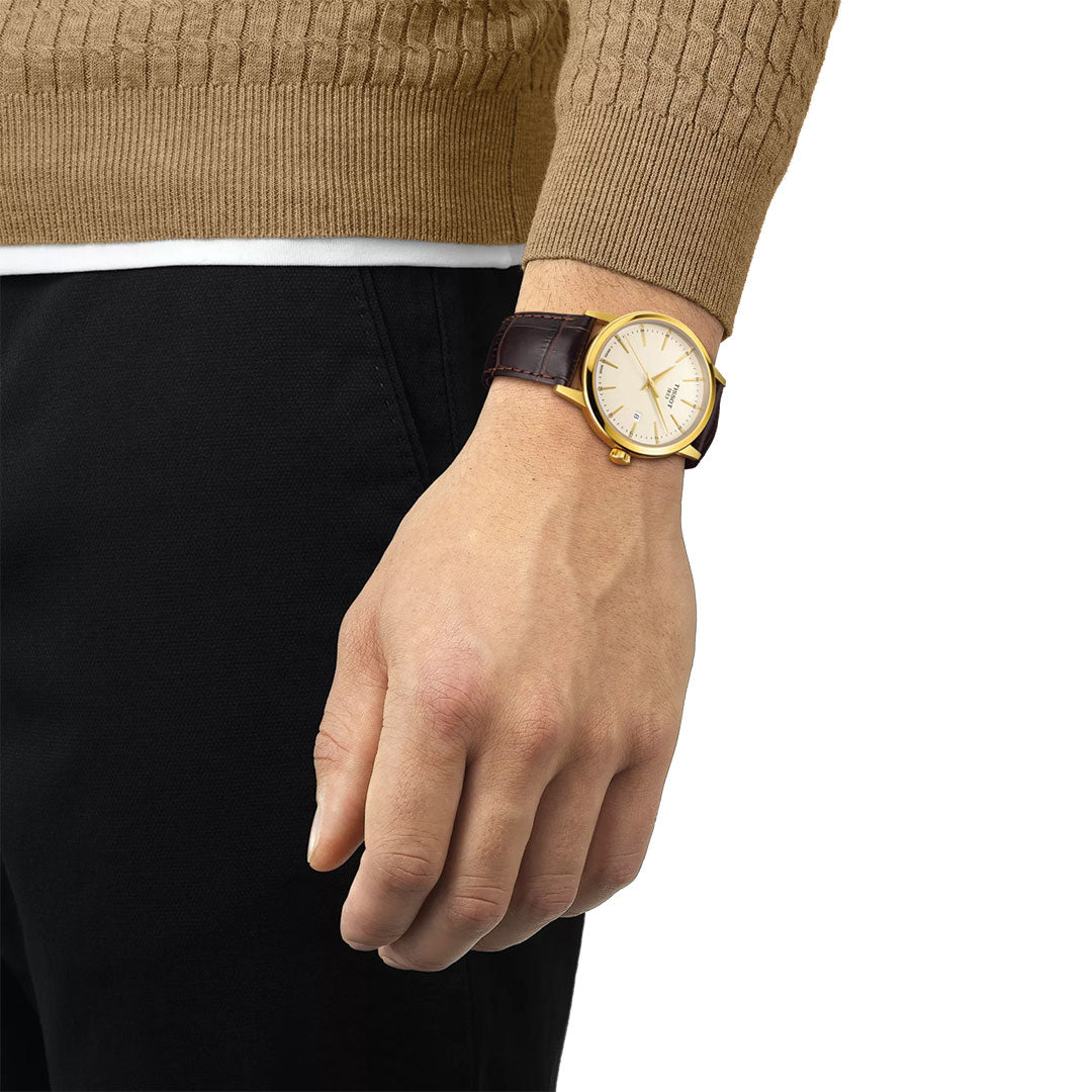 A person dressed in a tan sweater and black pants is shown from the waist down, wearing a Tissot Classic Dream Quartz 42mm Watch with a gold case and brown leather strap, their hand relaxed at their side.