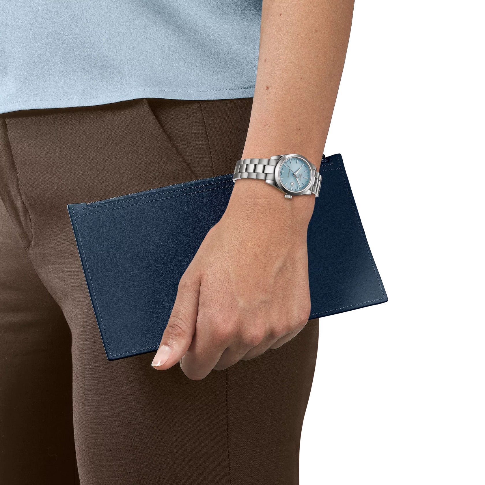 A person wearing a Tissot T-My Lady Automatic 29mm watch holds a navy blue leather wallet. They are dressed in a light blue top and brown pants, with the focus on the hand, wristwatch, and wallet against a plain background.