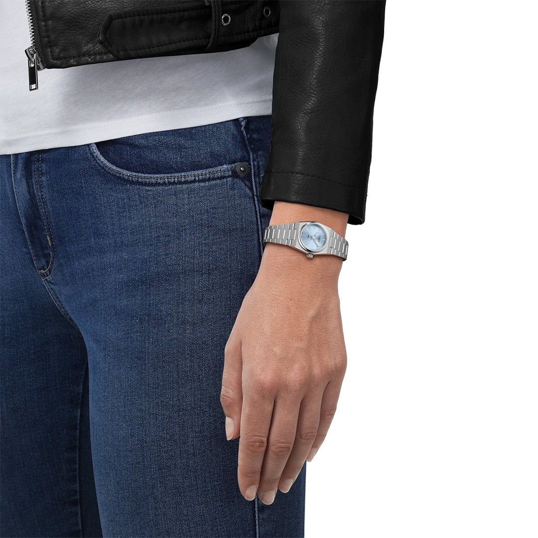 A person in a black leather jacket, white shirt, and blue jeans is shown from the waist down. Their left hand wears the Tissot PRX Quartz 25mm Watch with a light blue face, sapphire crystal, and stainless steel case by Tissot.