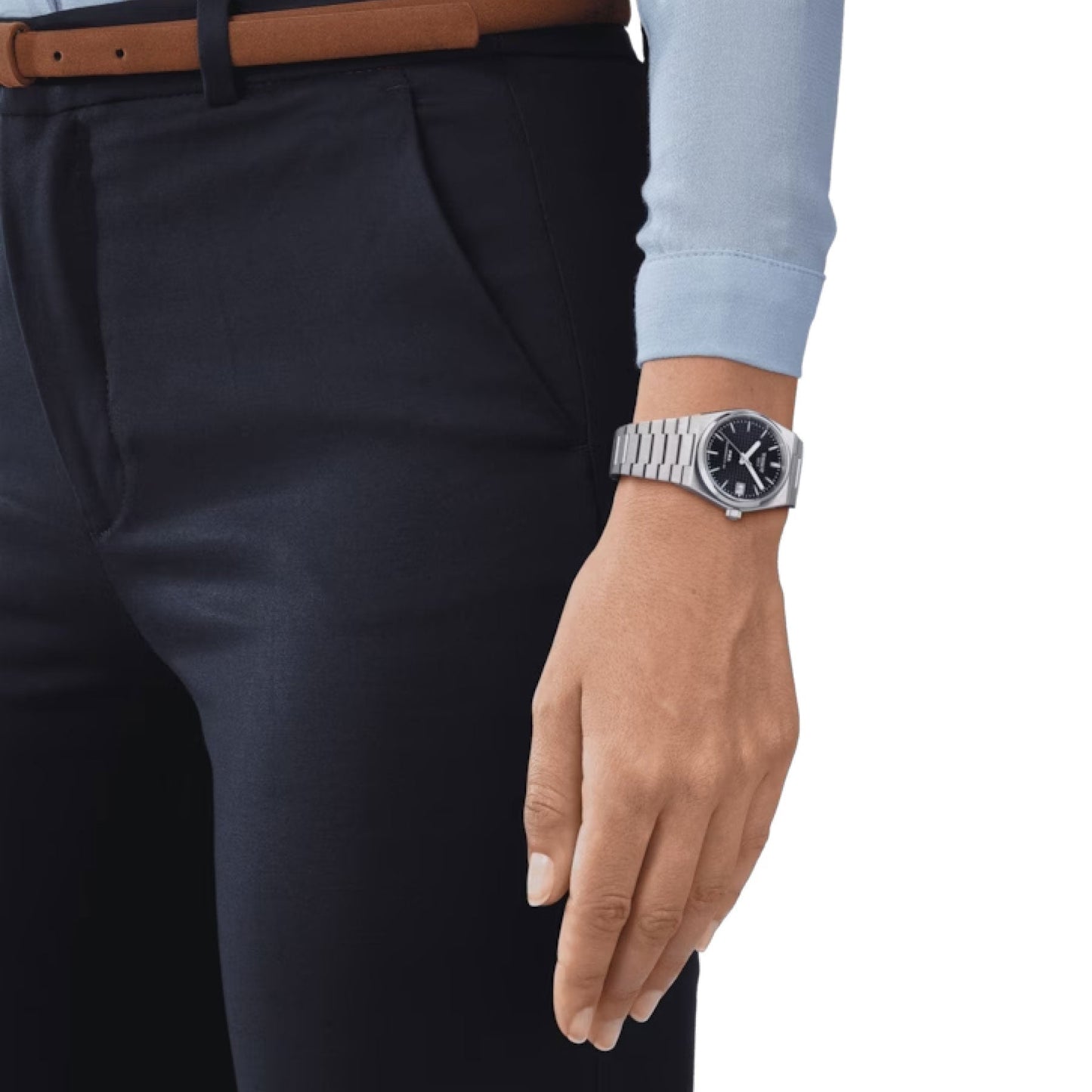 A person wearing a light blue long-sleeve shirt and dark pants is shown from the waist down. Their left arm is relaxed by their side, adorned with the Tissot PRX Powermatic 80 Automatic 35mm Watch featuring a 316L stainless steel case. A brown belt complements the outfit perfectly.