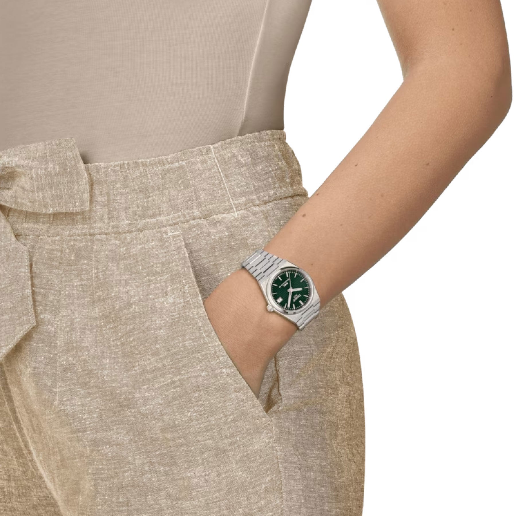 A person wearing light beige pants with a tied belt and matching top has their hand in the pocket, showcasing a stylish Tissot PRX Powermatic 80 Automatic 35mm Watch. The timepiece features a dark green face with Swiss automatic movement, elegantly highlighted against the plain white background.