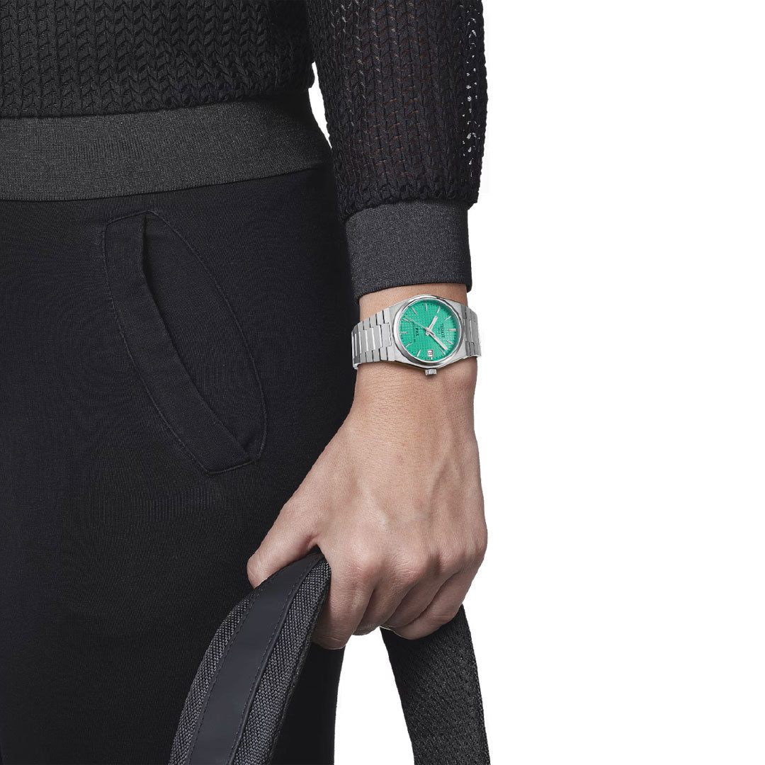 A person is wearing a Tissot PRX Powermatic 80 Automatic 35mm Watch with a silver band and green face that features an interchangeable quick release bracelet. They are holding a black textured item, likely a bag, while dressed in a black and gray ensemble that includes a textured sweater and pants with visible pockets. The emphasis is on the watch and their hand.