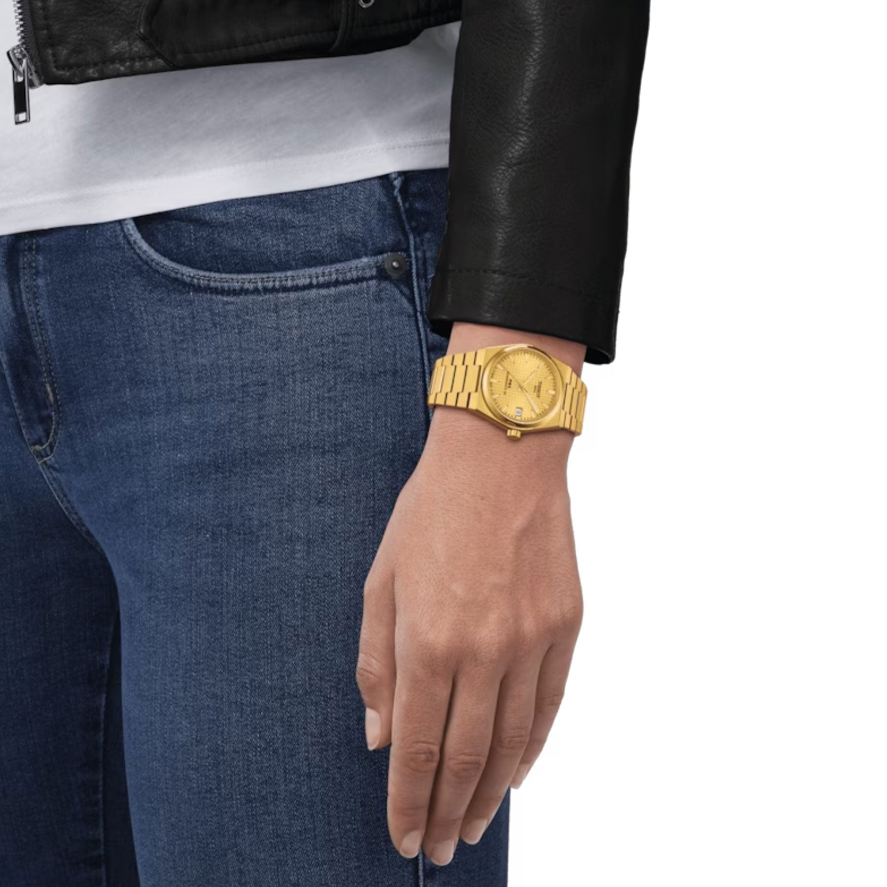 A person clad in a black leather jacket and blue jeans is depicted from the waist down, with their left hand gently resting by their side. The wristwatch they are wearing is a Tissot PRX Powermatic 80 Automatic 35mm Watch, showcasing a yellow gold PVD-coated linked bracelet. The backdrop is a simple white setting.