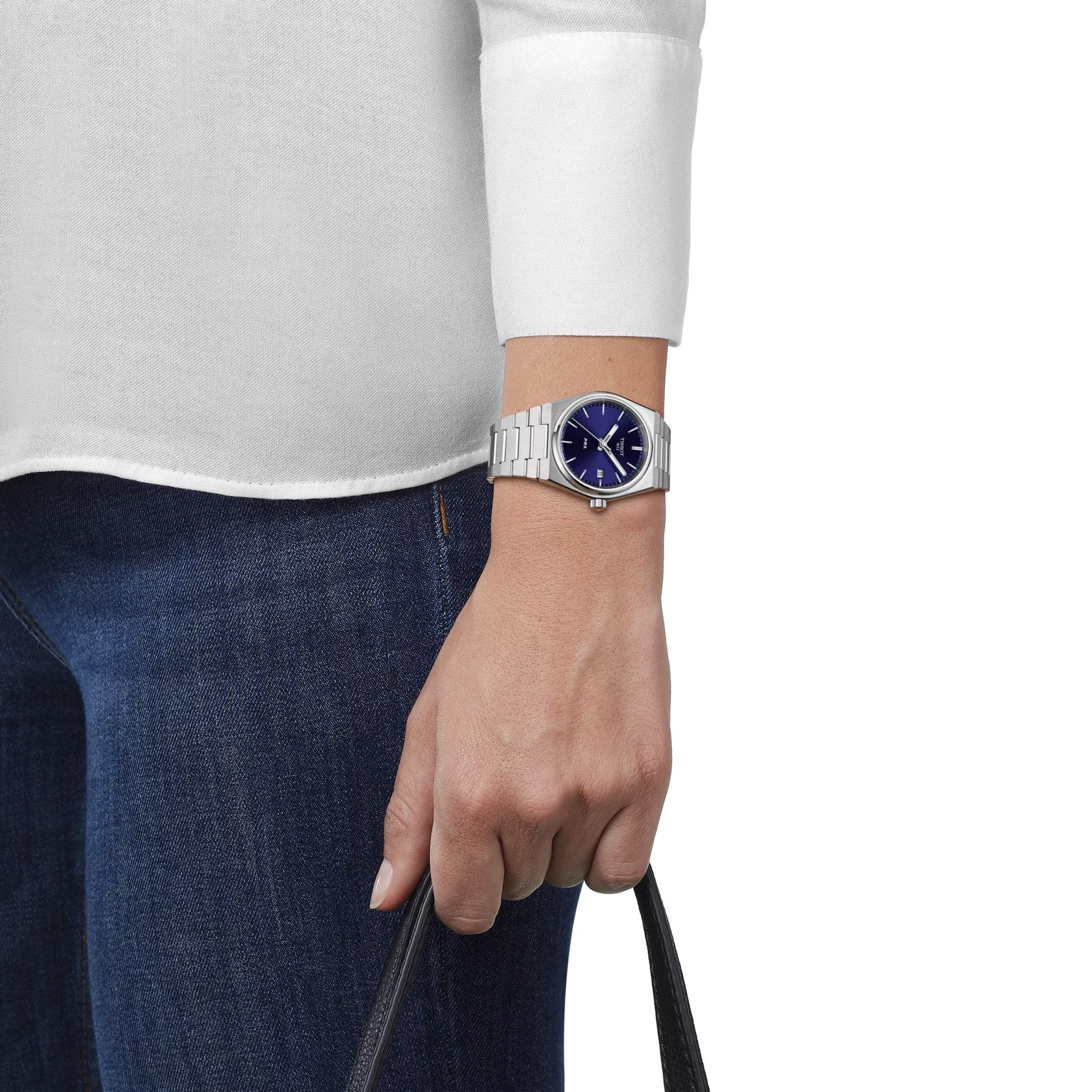 A person in a white long-sleeve shirt and blue jeans holds a black handbag, wearing a Tissot PRX Quartz 35mm watch featuring a retro design, silver case with blue dial, and sleek stainless steel integrated bracelet.