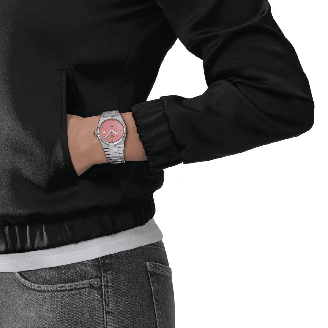 A close-up of an individual dressed in a black jacket and jeans with one hand in their pocket highlights the chic Tissot PRX Quartz 35mm Watch. The watch features a stylish silver finish and a pink face, complete with Swiss Quartz movement and a robust stainless steel case, standing out against the plain white background.