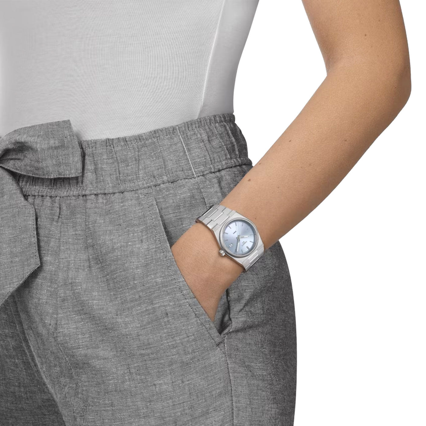 A person wearing grey pants with a bow detail and a white top has their hand in their pocket. They sport a silver Tissot PRX Quartz 35mm watch featuring a sapphire crystal and Swiss quartz movement, complete with a blue face.