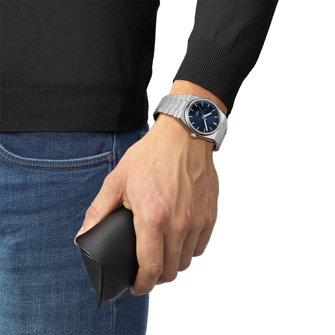 Wearing a Tissot PRX UFO Robot Grendizer Powermatic 80 Automatic 40mm Watch and a black long-sleeve top, a person holds a black leather glasses case against their blue jeans.