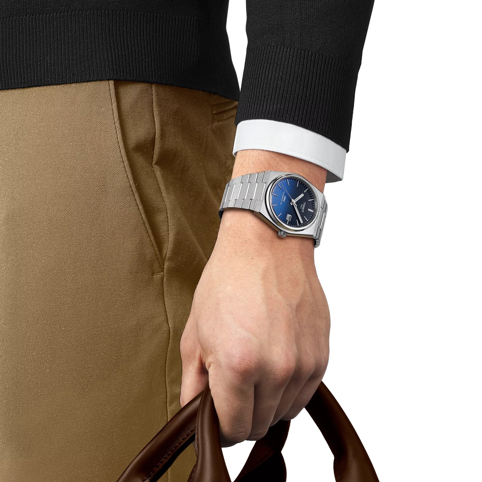 An individual sporting a Tissot PRX Gradient Powermatic 80 Automatic 40mm watch, which boasts a blue gradient dial with sapphire crystal and a metallic band. They are clad in a black sweater with white cuffs, carrying a brown leather bag. Their brown pants serve as a contrast to the watch's hue.