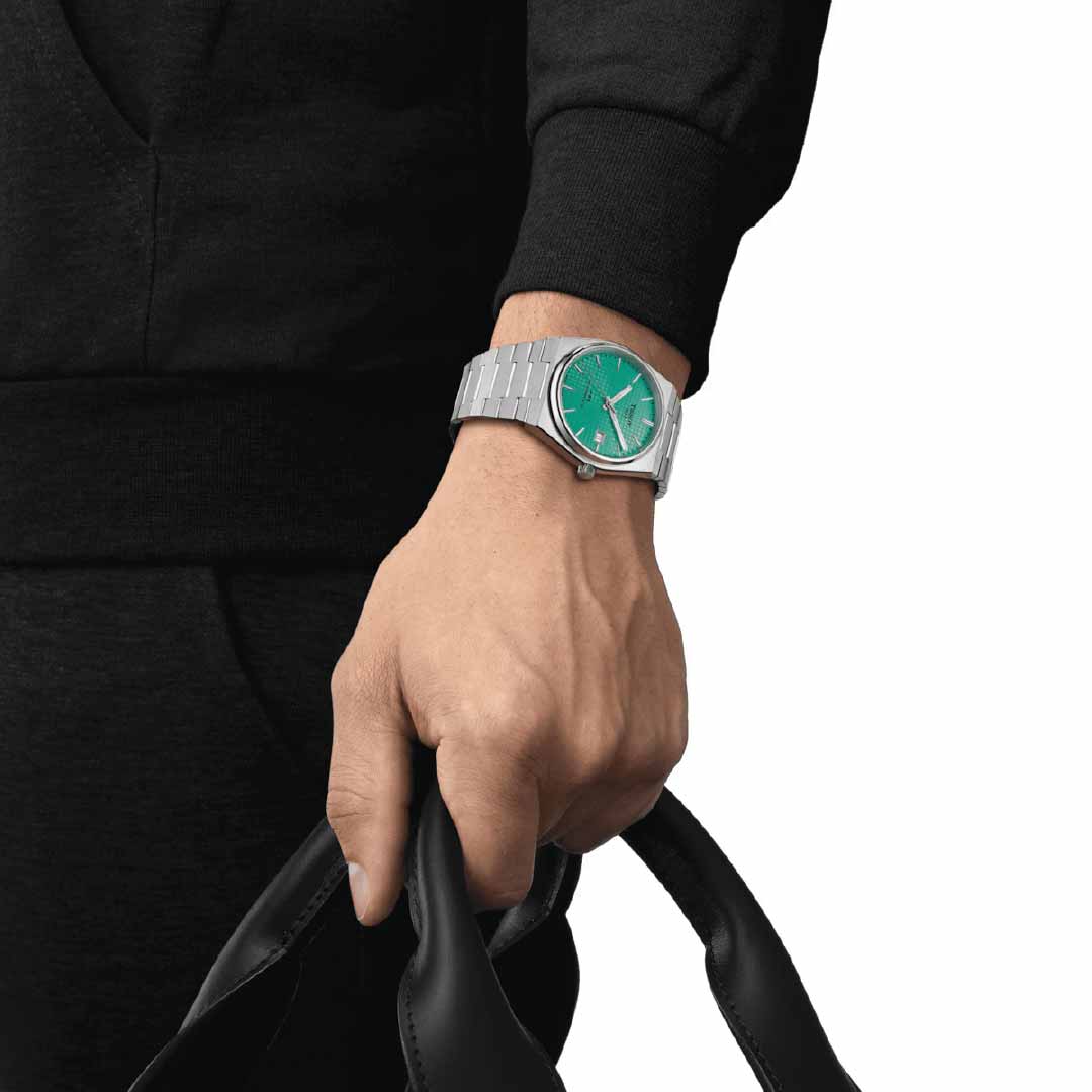 The individual, dressed in black clothing, is captured from the chest down against a plain white background, wearing a Tissot PRX Powermatic 80 Automatic 40mm Watch with a silver finish and scratch-resistant sapphire crystal featuring a green face, while holding a black leather bag.