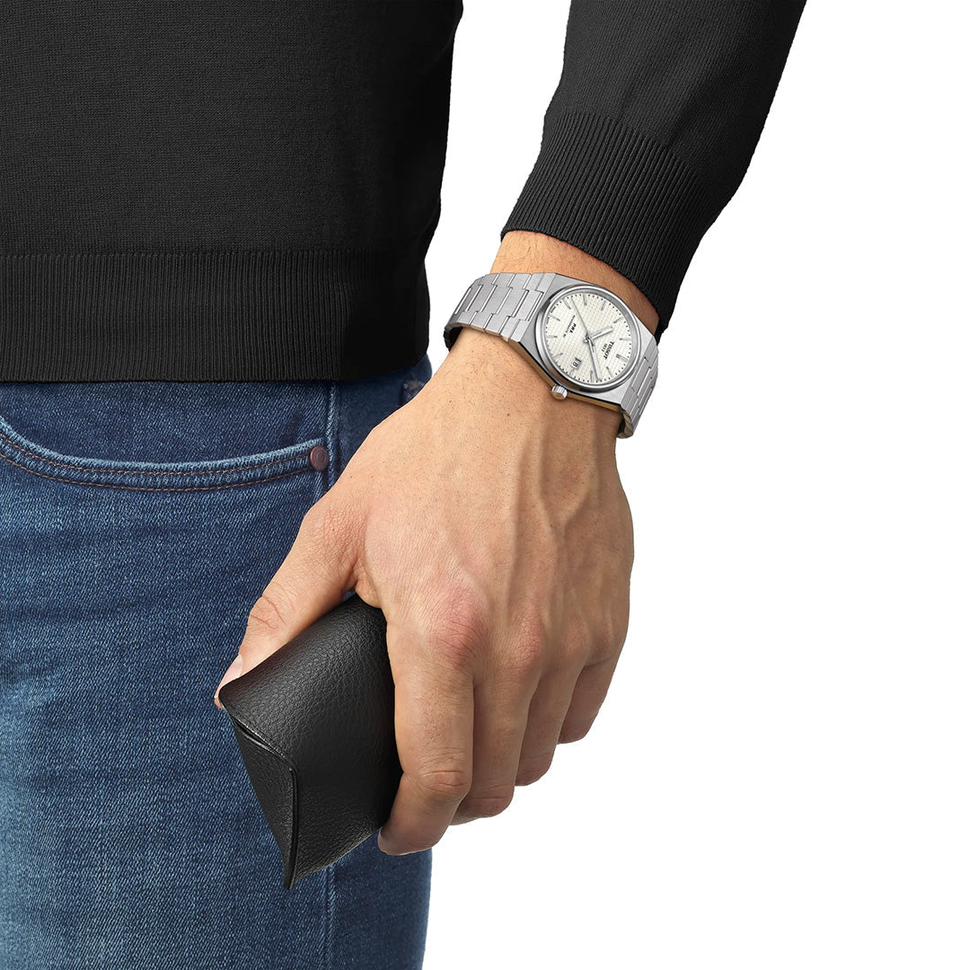 A person wearing a Tissot PRX Powermatic 80 Automatic 40mm watch with a silver finish and a Nivachron hairspring, paired with a black sweater, holds a black eyeglass case in their right hand against a plain background.