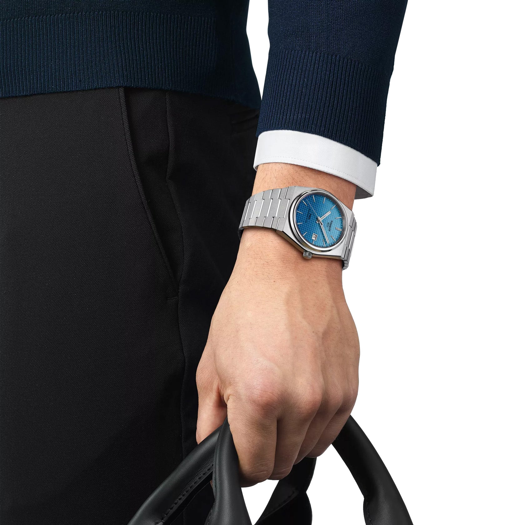 A close-up shows a hand wearing a Tissot PRX Powermatic 80 Automatic 40mm Watch with a blue gradient dial and sapphire crystal, holding a black leather bag. The person is dressed in a dark sweater over a white shirt.