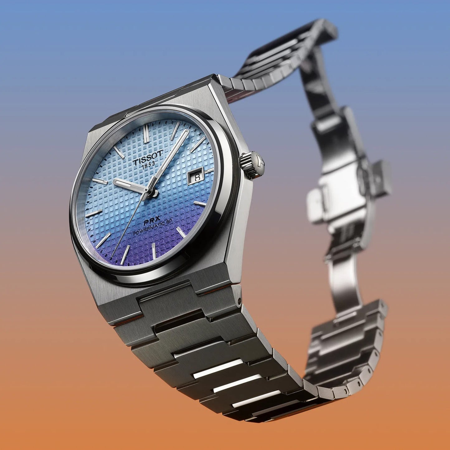 The Tissot PRX Powermatic 80 Automatic 40mm Watch boasts a blue textured gradient dial and silver metal strap, accented by a vibrant blue-to-orange background. It features hour markers, hands, a date window, sapphire crystal, and the distinguished Tissot logo on the dial.