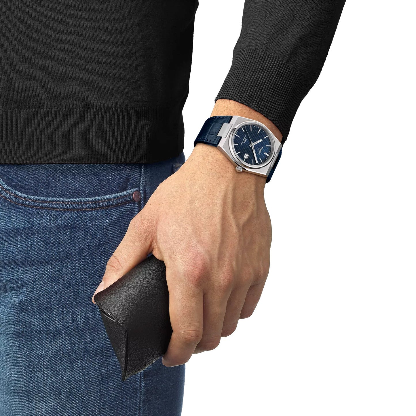 A person in a dark sweater and jeans holds a black leather wallet, wearing the stylish Tissot PRX Powermatic 80 Automatic 40mm Watch featuring a blue face, silver frame, stainless steel case, classic design with hour markers, date display, and impressive power reserve for reliability.