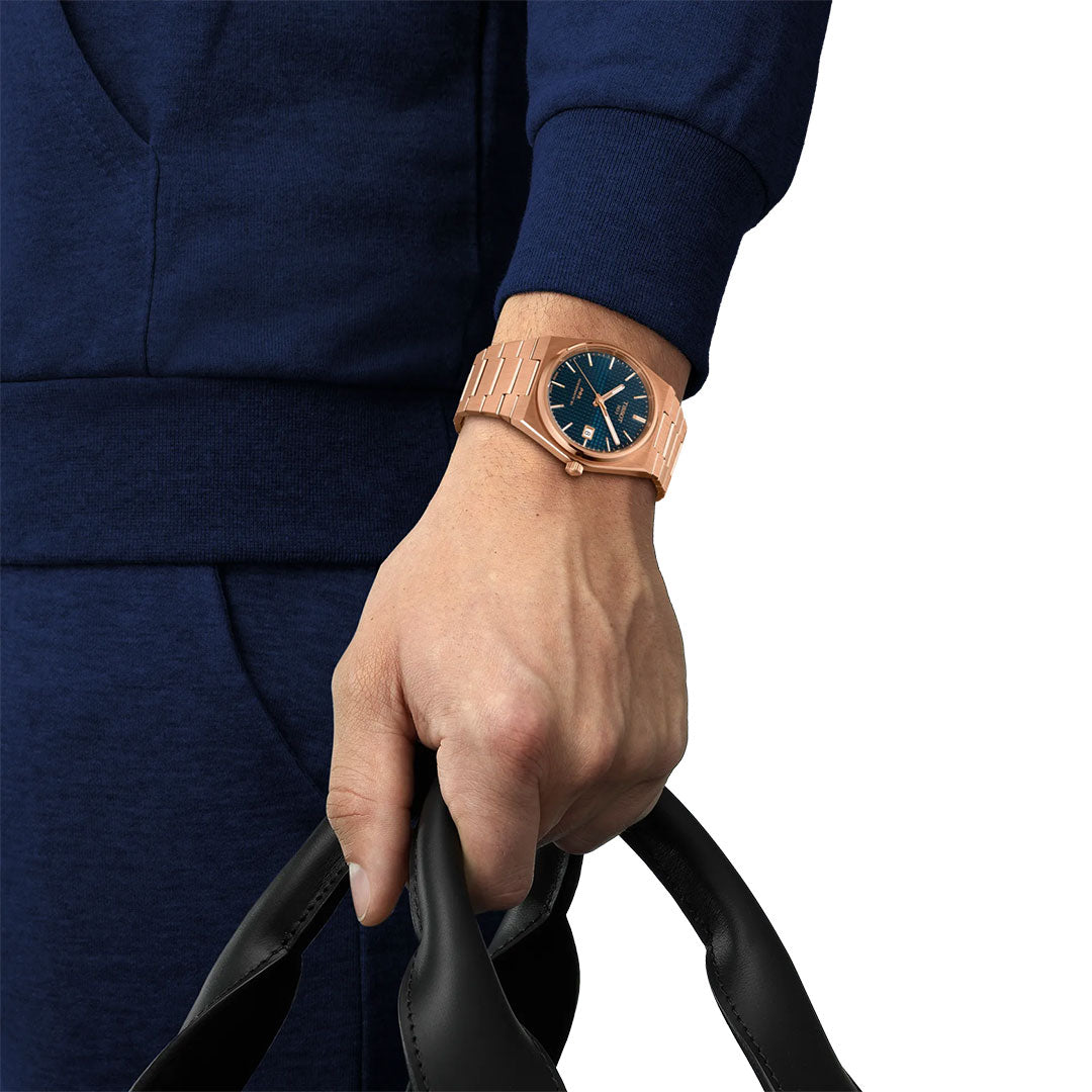 Against a white background, a person in blue holds black leather handles while wearing a gold Tissot PRX Powermatic 80 Automatic 40mm Watch with a dark blue face.