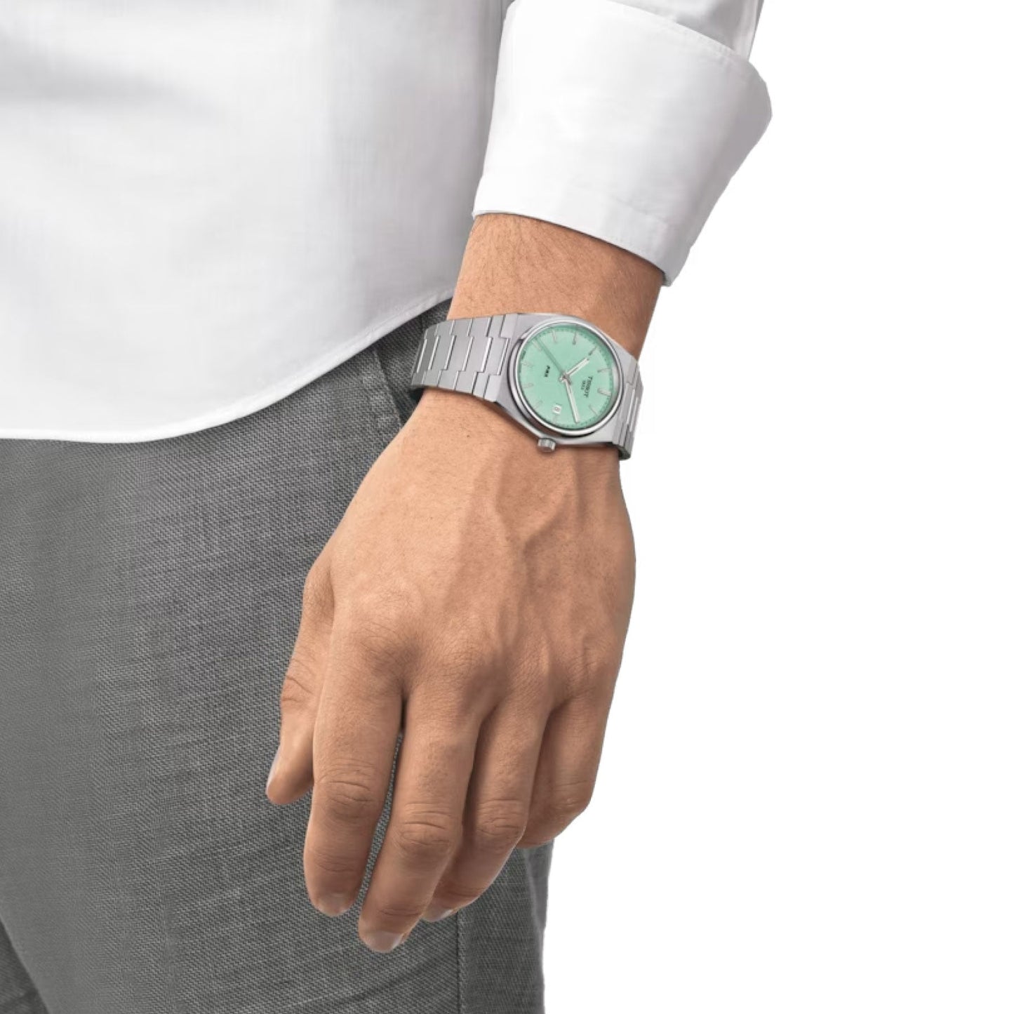 A person clad in a white shirt and gray pants is depicted with a Tissot PRX Quartz 40mm Watch, which boasts a sleek silver design complemented by a light green face encased in stainless steel, on their left wrist. The hand, adorned with an interchangeable bracelet, rests calmly at their side against a white backdrop.