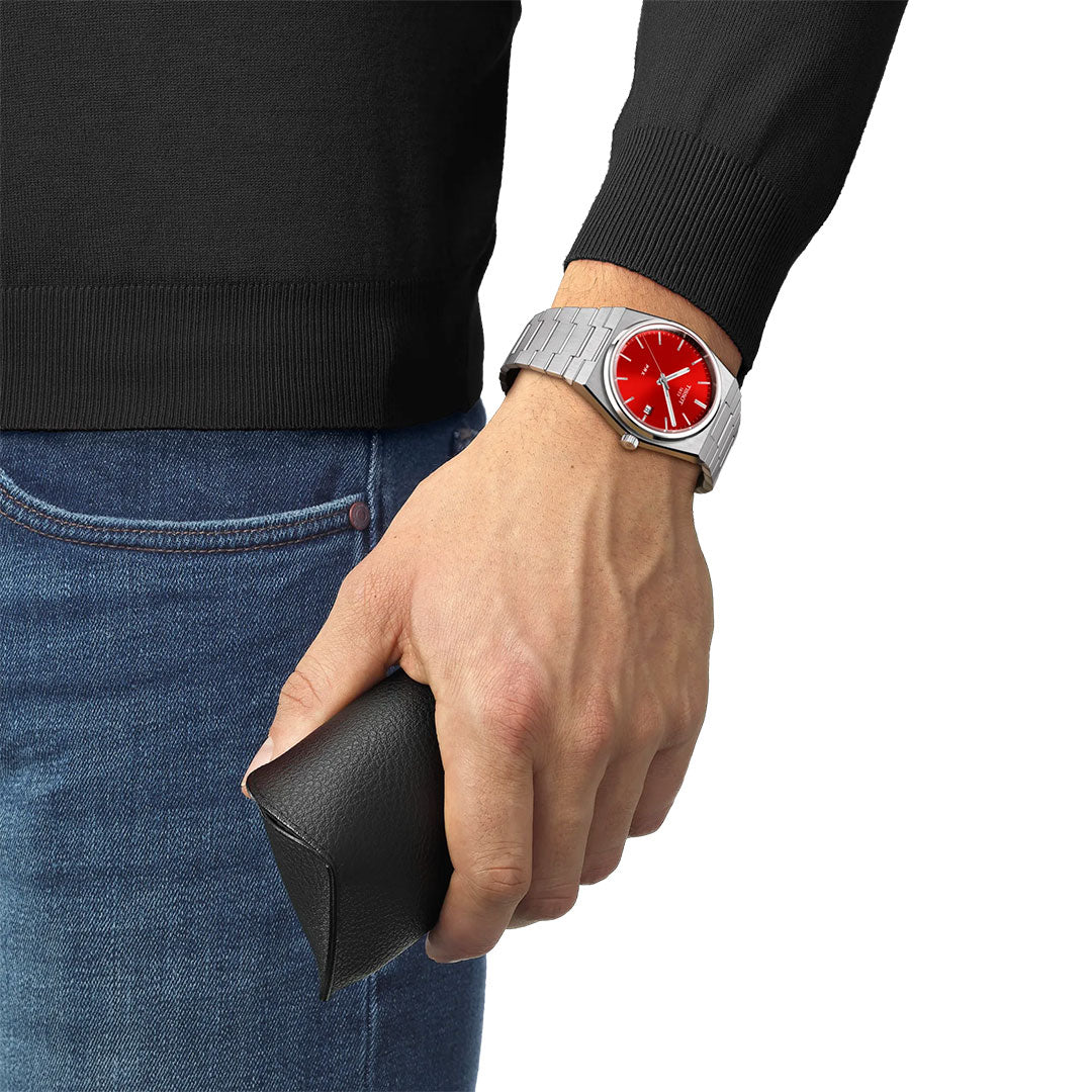 A person in a black sweater and blue jeans holds a black eyeglass case and wears a Tissot PRX Quartz 40mm Watch by Tissot, featuring a silver band and striking red face—ideal for design lovers.