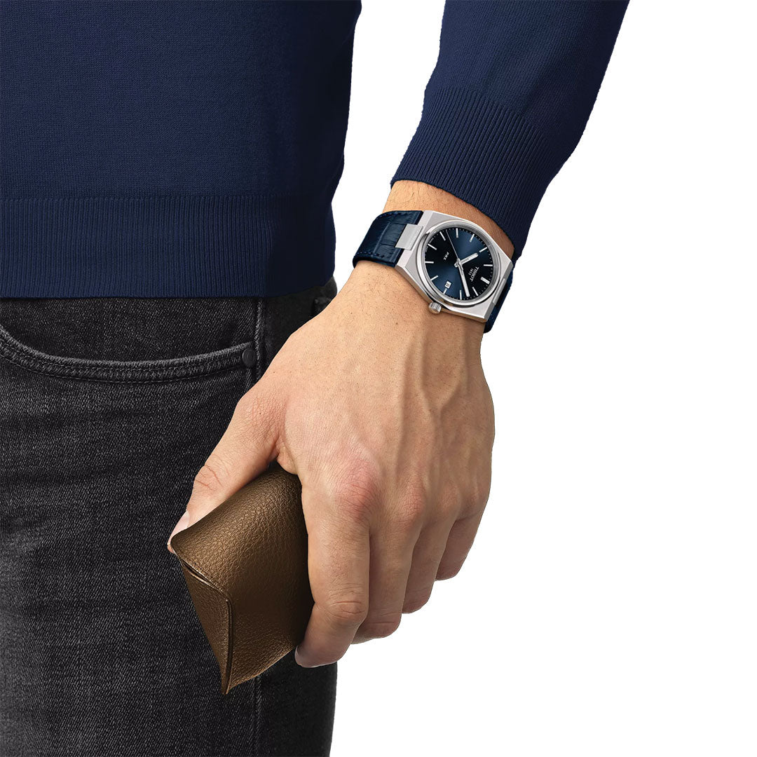 A person in a blue sweater and black jeans holds a brown leather pouch, showcasing the Tissot PRX Quartz 40mm Watch by Tissot, featuring a silver 316L stainless steel case with a blue dial and matching blue strap.