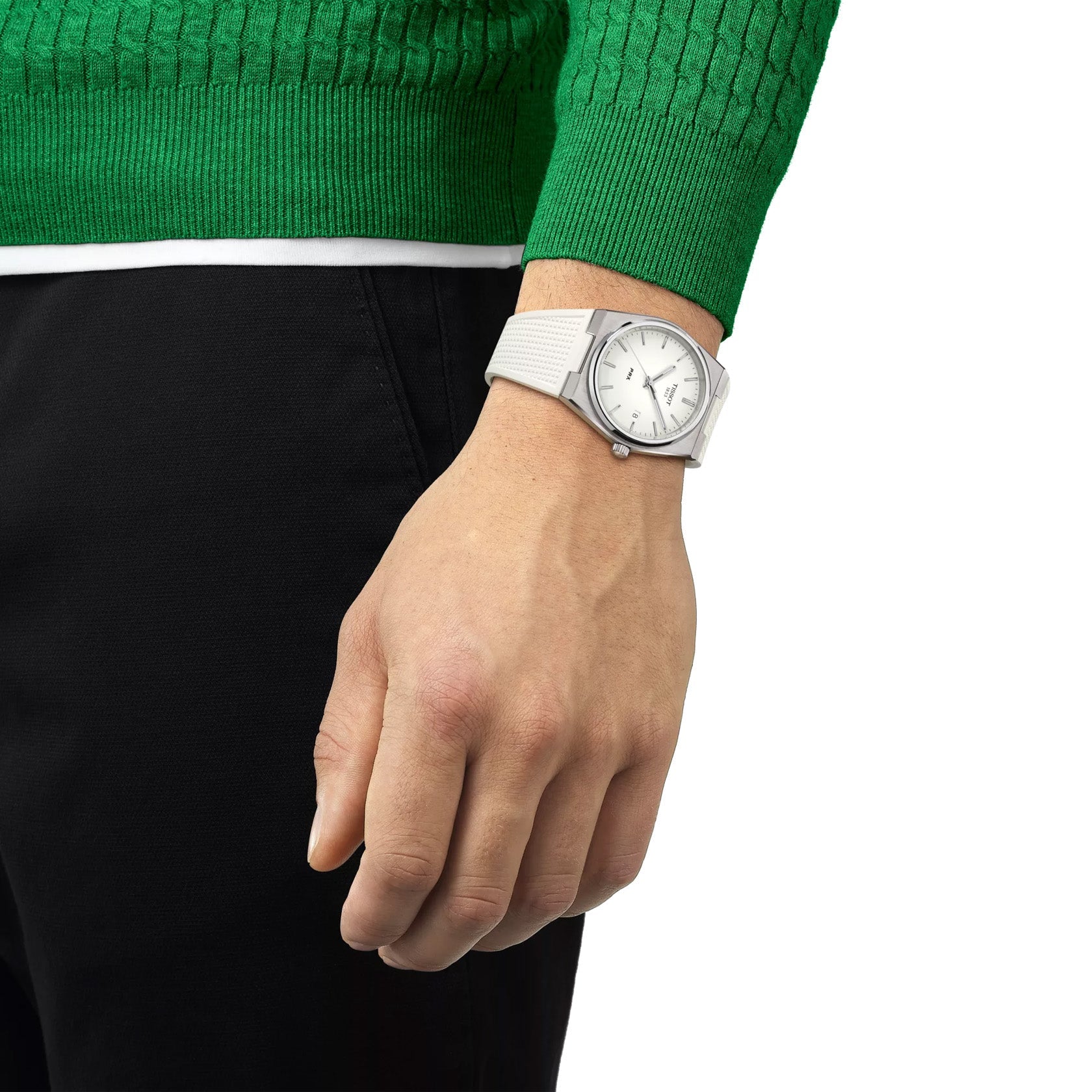 A person in a green sweater and black pants showcases the Tissot PRX Quartz 40mm wristwatch with modern-vintage flair, featuring a slim design, white band, and watch face. The stylish watch adorns the left wrist as the arm rests downward.