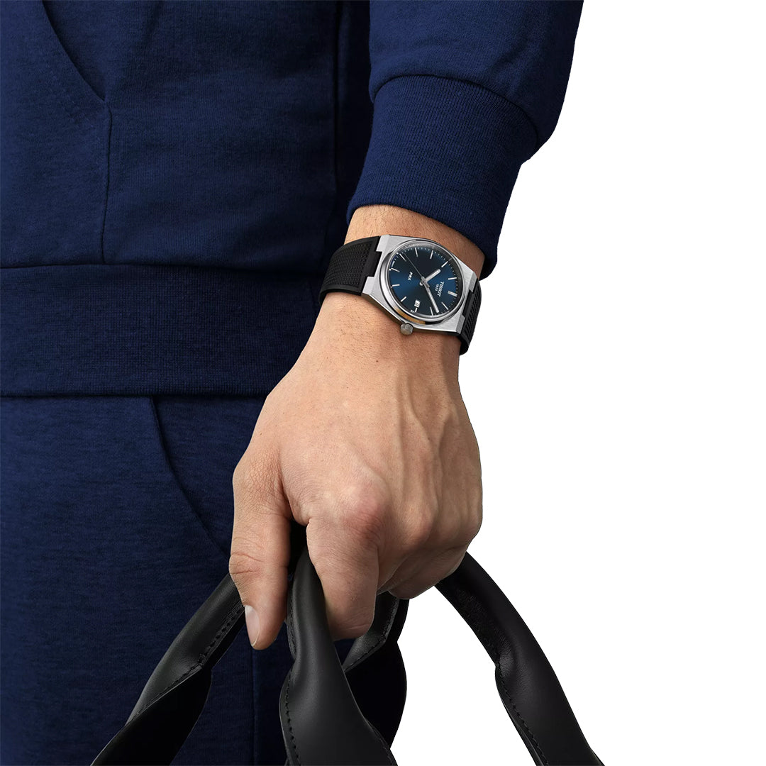 A person in blue holds black handles, wearing a Tissot PRX Quartz 40mm Watch—a silver Tissot wristwatch with a blue dial and black strap—on their left wrist.