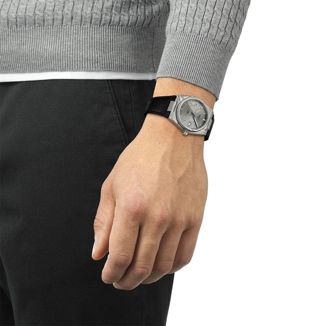 A person in a gray sweater and black pants wears a Tissot PRX Damascus Steel Powermatic 80 Automatic 38mm Watch with a black band on their left wrist, the marbled Damascus steel case catching the light as their hand rests at their side.