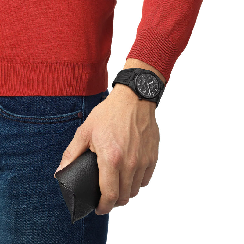 A person wearing a red sweater and blue jeans holds a black case in their hand, showcasing the Tissot PRX Carbon Powermatic 80 Automatic 40mm Watch, featuring a round face with sapphire crystal. The image emphasizes the person's hand and the items they are holding.