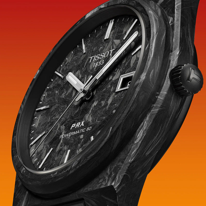 Close-up of a Tissot PRX Carbon Powermatic 80 Automatic 40mm Watch, featuring a black marbled dial with white hour markers and silver hands, housed in a forged carbon case. The watch face displays the date and the Tissot brand name against an orange gradient background, all safeguarded by sapphire crystal.