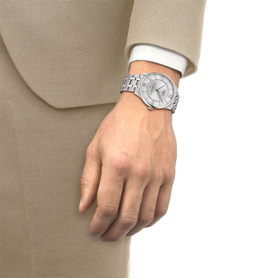 A person in a beige suit and white shirt wears a Tissot Chemin Des Tourelles Powermatic 80 Automatic 42mm Watch with a sapphire crystal and metal band on their left wrist, its light silver dial visible as their relaxed arm rests by their side.