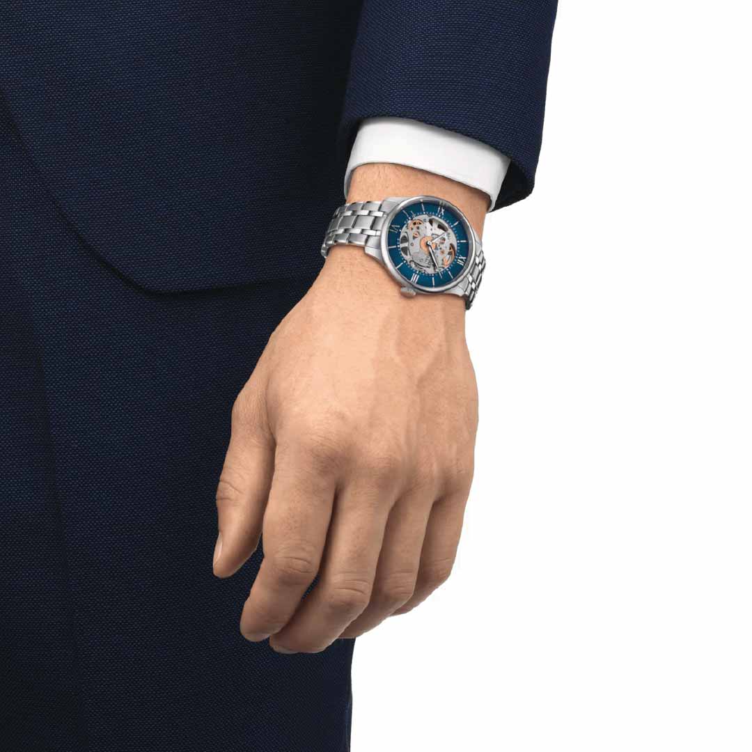 A close-up of a person wearing a dark blue suit, highlighting their wrist adorned with the Tissot Chemin des Tourelles Skeleton Automatic 39mm Watch. This sophisticated timepiece, featuring Swiss automatic movement and a silver and blue design, complements their relaxed hand positioned at their side against a plain white background.