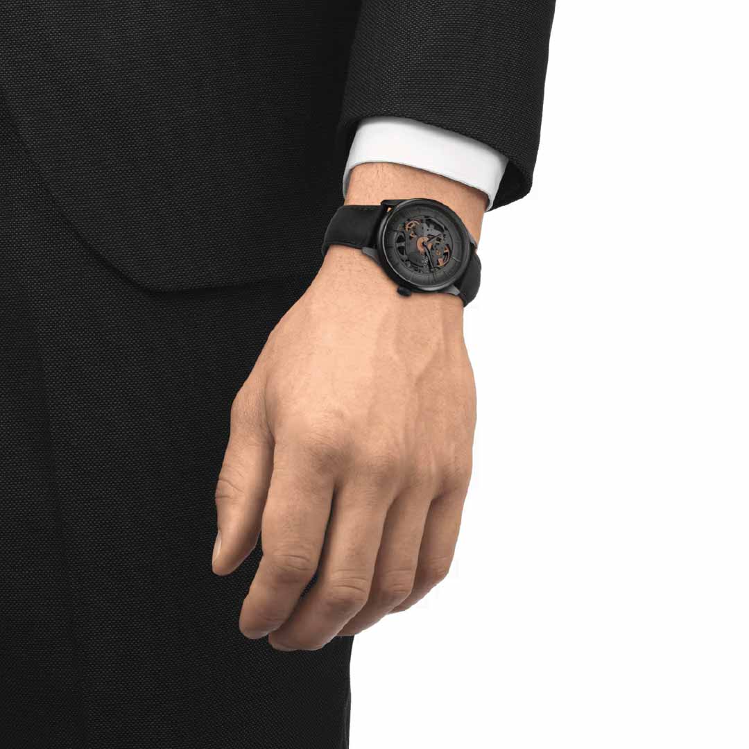 A close-up of a person's left hand wearing a black suit showcases the Tissot Chemin des Tourelles Skeleton Automatic 39mm Watch, highlighted by its Swiss automatic movement and exposed mechanical design. The scratch-resistant sapphire crystal adds sophistication to the formal attire, completing the elegant look with refined luxury from Tissot.