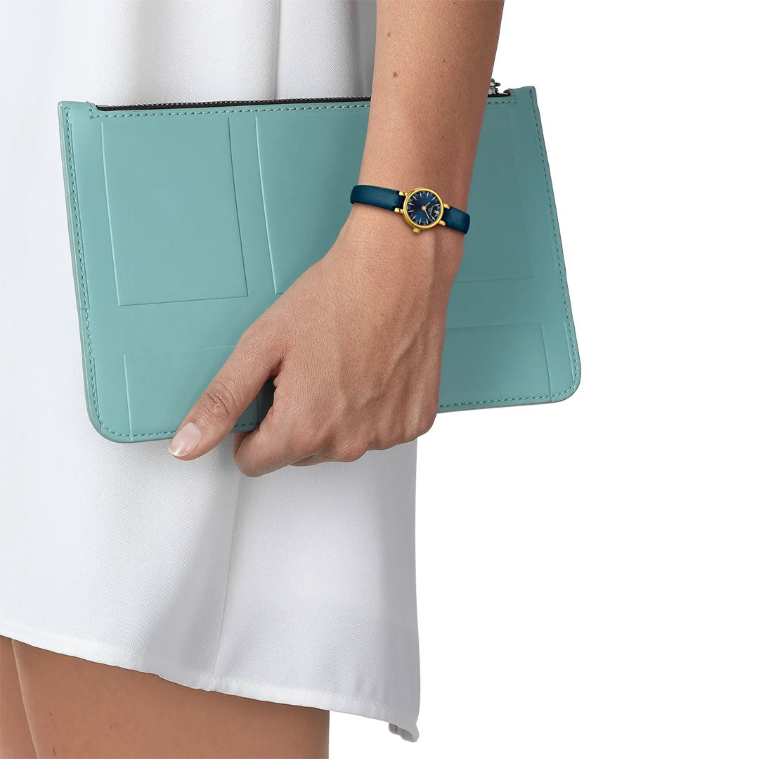 Wearing a white skirt, a person holds a light blue clutch and sports the Tissot Lovely Round Quartz 19.5mm Watch with a blue dial and gold accents by Tissot on their left wrist.