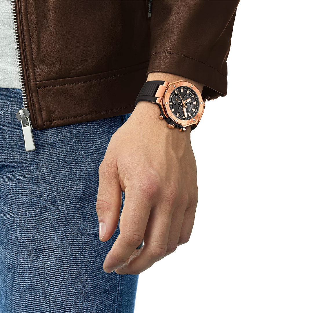 A person in a brown leather jacket and blue jeans wears a Tissot T-Race Chronograph Quartz 45mm Watch by Tissot with a black band on their left wrist, their hand relaxed and hanging at their side.