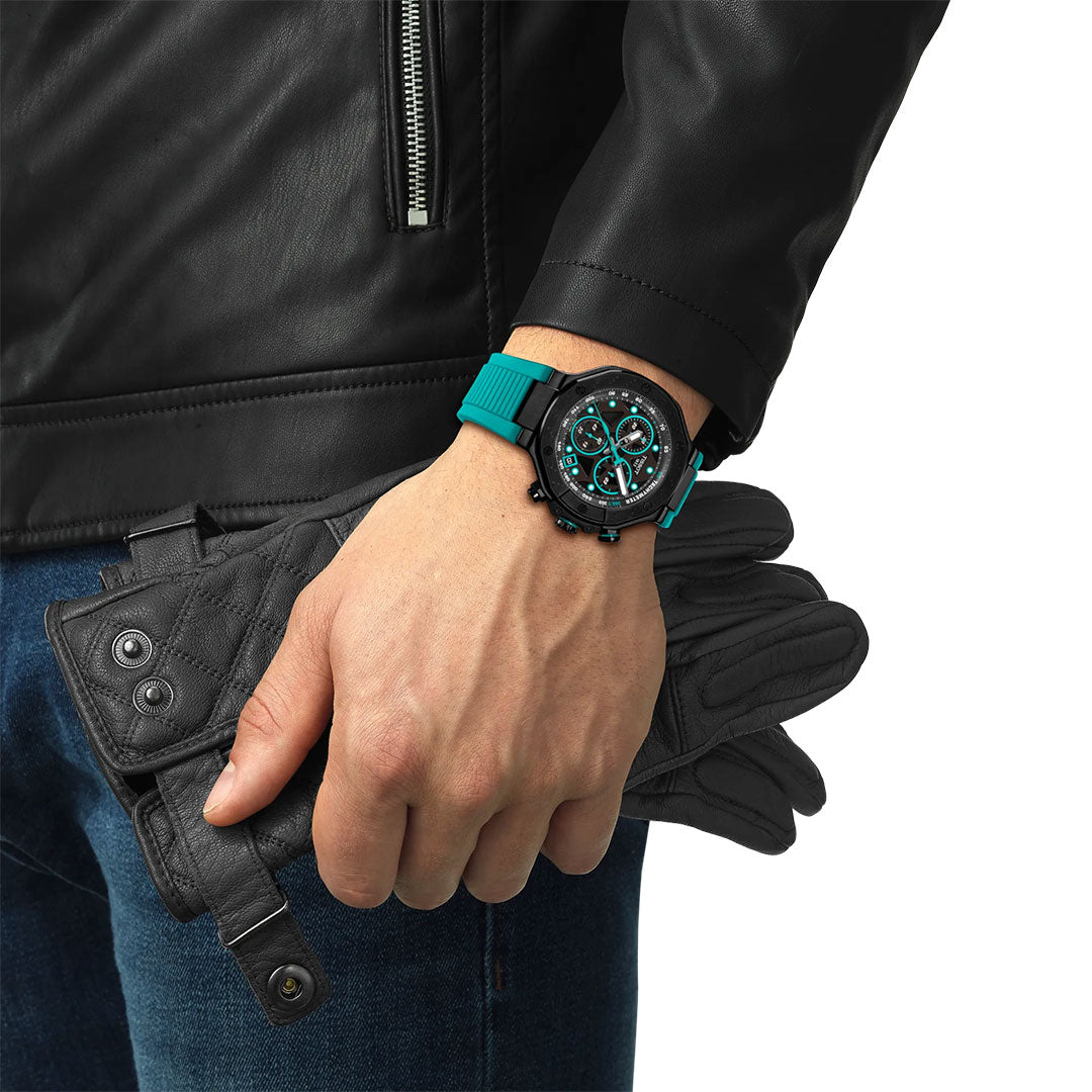 A person in a black leather jacket and jeans holds black leather gloves, showing off the Tissot T-Race Chronograph Quartz 45mm Watch with a teal strap—an accessory by Tissot inspired by racing bikes.
