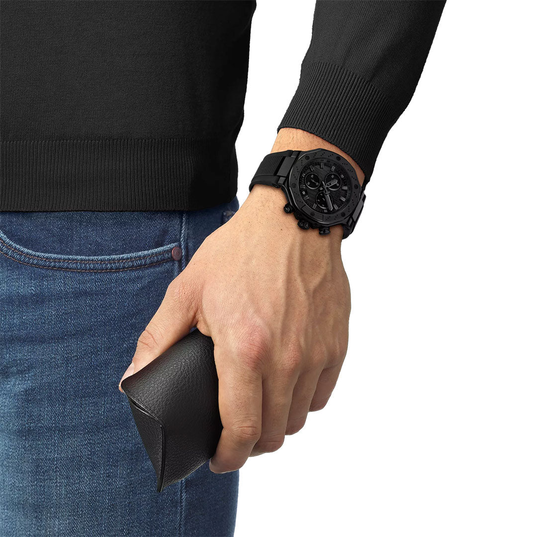 A person in a black long-sleeve shirt, blue jeans, and a Tissot T-Race Chronograph Quartz 45mm Watch by Tissot holds a black textured eyeglass case in their right hand.