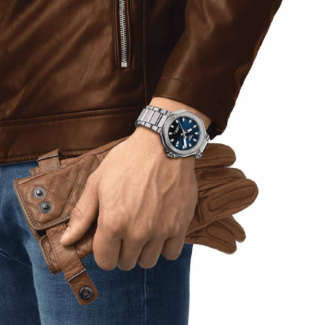 A person dressed in a brown leather jacket and blue jeans holds a pair of brown leather gloves, embodying the rugged spirit of motorbikes. On their wrist, the Tissot T-Race Powermatic 80 Automatic 41mm Watch gleams with its blue face against the plain white background.