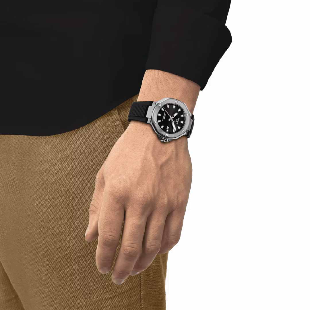 A person is wearing the Tissot T-Race Powermatic 80 Automatic 41mm Watch, featuring a silver bezel and a black strap. The Powermatic 80 mechanism presents time on an elegant black dial. Dressed in a black shirt and brown pants, they stand with one hand hanging relaxed by their side, exuding effortless style.