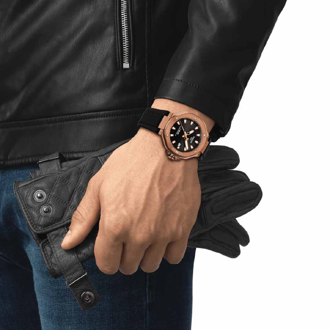 A person in a black leather jacket and blue jeans holds black leather gloves. On their left wrist, they sport a Tissot T-Race Powermatic 80 Automatic 41mm watch, known for its style and precision, showcased against a plain white background.