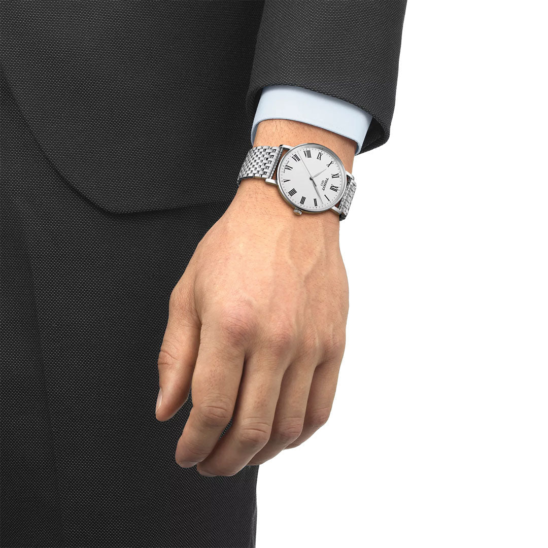 A person in a dark suit and light blue shirt wears a Tissot Everytime Quartz 40mm Watch with a white face, Roman numeral markers, and stainless steel bracelet on their left wrist.