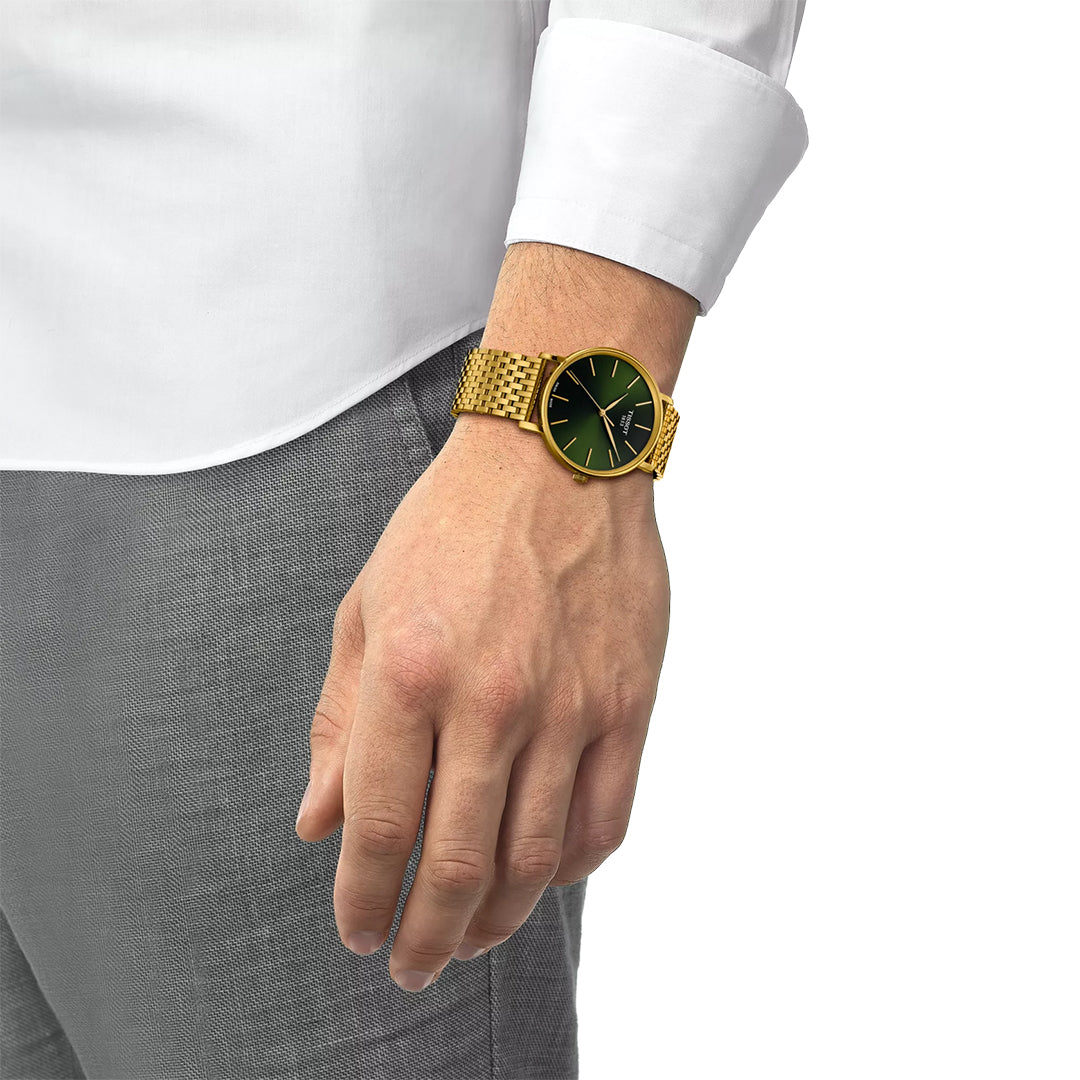 A person in a white shirt and gray pants wears a Tissot Everytime Quartz 40mm Watch with a gold case, green dial, and stainless steel details on their left wrist against a plain white background.