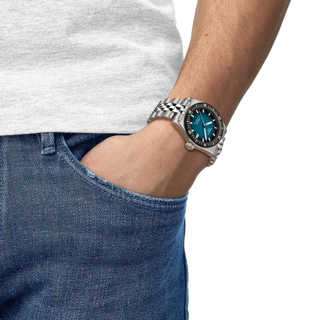 A person wears a Tissot PR516 Powermatic 80 Automatic 38mm stainless-steel watch with a teal face and metal band, hand in blue jeans pocket, paired with a white t-shirt. The Tissot timepiece is prominently displayed.