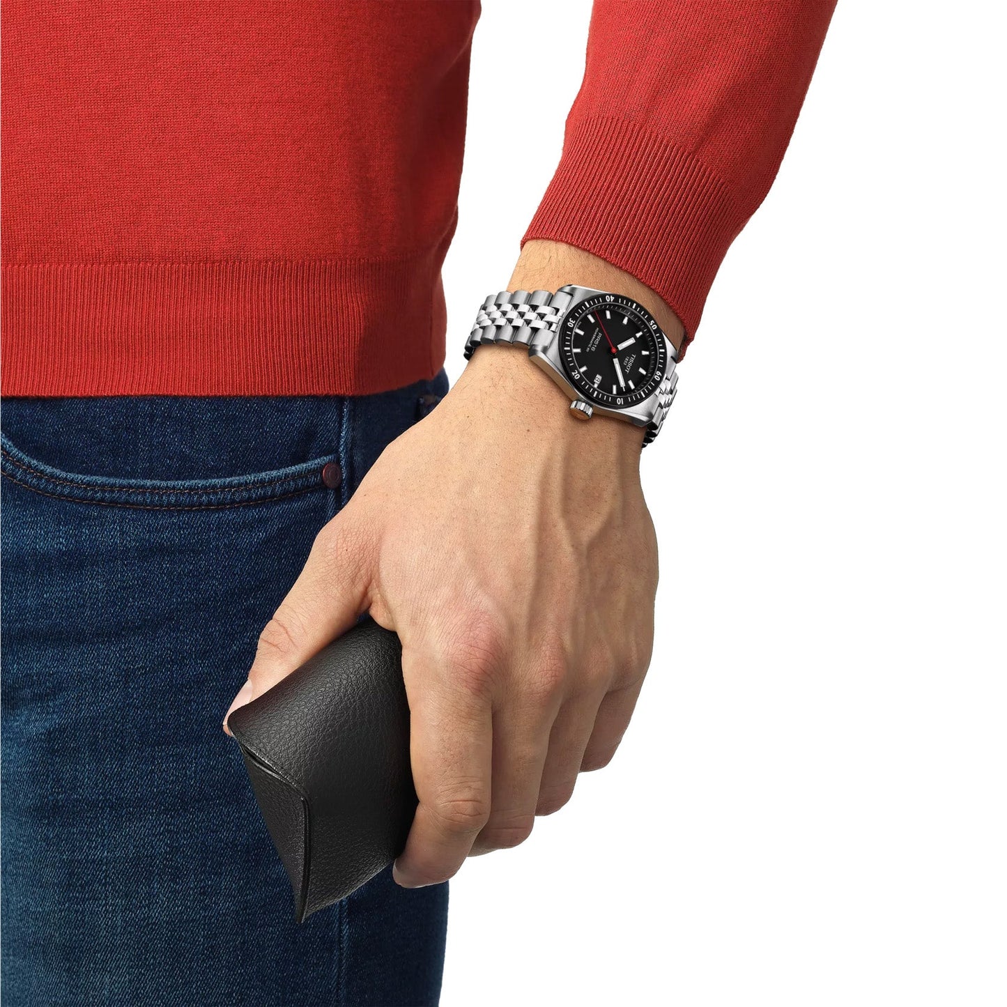 A person in a red sweater holds a black sunglasses case in their right hand. Their wrist is adorned with a Tissot PR516 Powermatic 80 Automatic 38mm Watch featuring a stainless-steel case. They wear blue jeans, and only their torso and hands are visible.