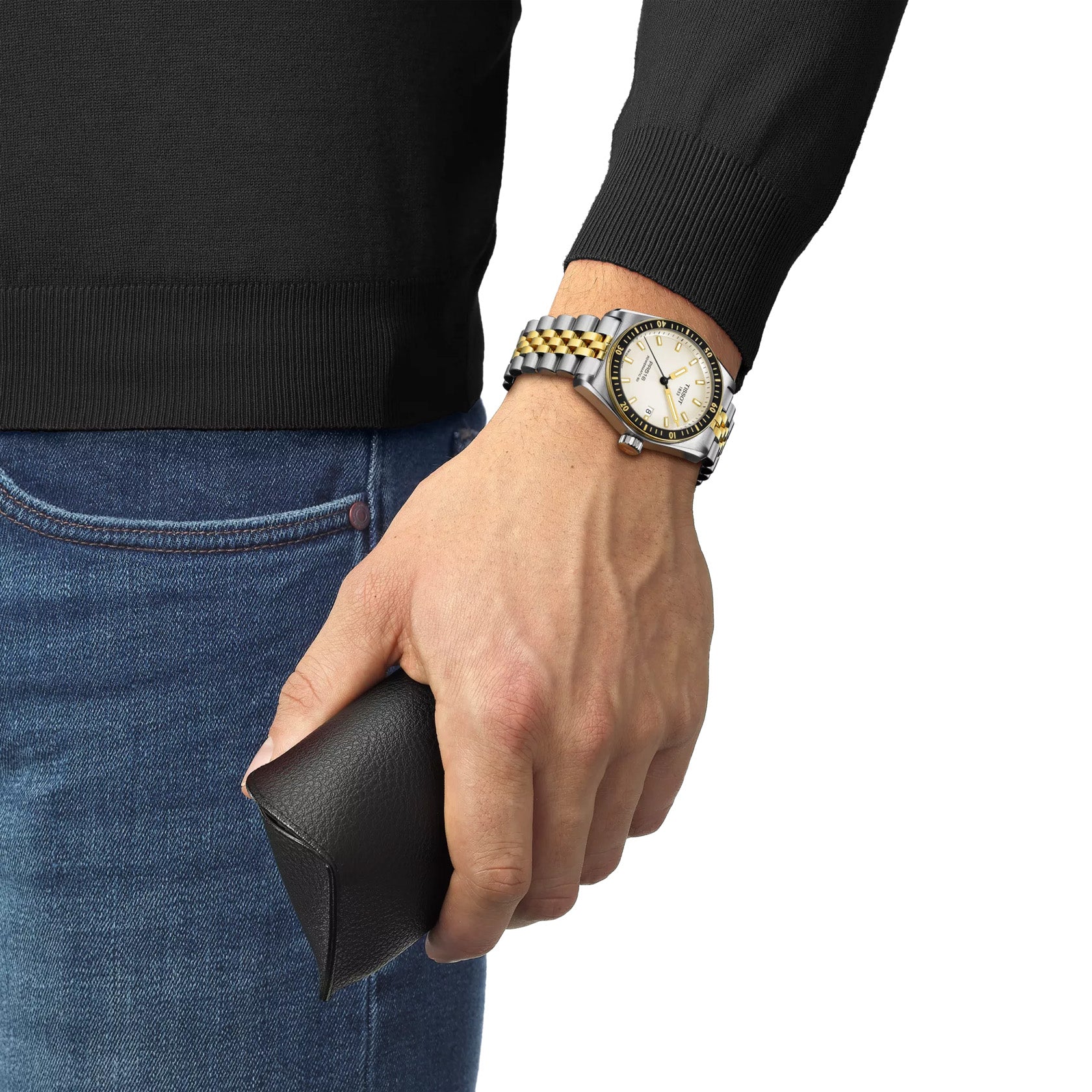 A person in a black sweater and jeans holds a sleek black leather wallet. Their wrist features the Tissot PR516 Powermatic 80 Automatic Watch, boasting Super-LumiNova®️ indices within a 38mm stainless-steel case. The plain white background enhances their understated elegance.