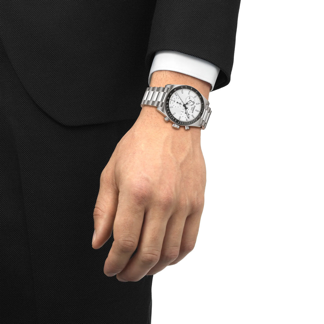 A person in a black suit and white shirt wears a Tissot PR516 Chronograph Quartz 40mm Watch by Tissot with a stainless-steel bracelet on their left wrist. Only the lower arm and hand are visible.