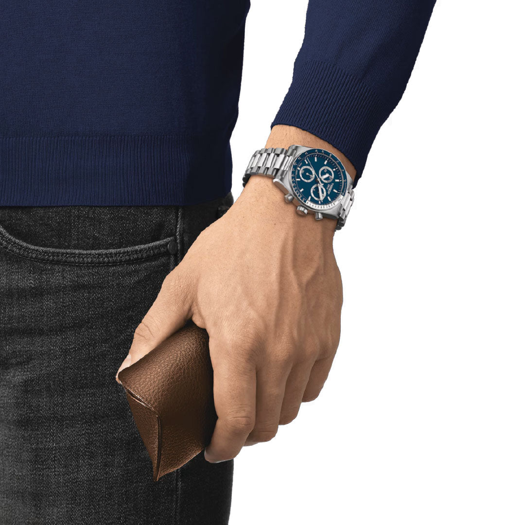 A person wearing a blue sweater and dark jeans holds a brown leather wallet in their right hand. On their left wrist, they sport the sleek Tissot PR516 Chronograph Quartz 40mm Watch, which showcases its striking blue face and adds elegance to the ensemble.